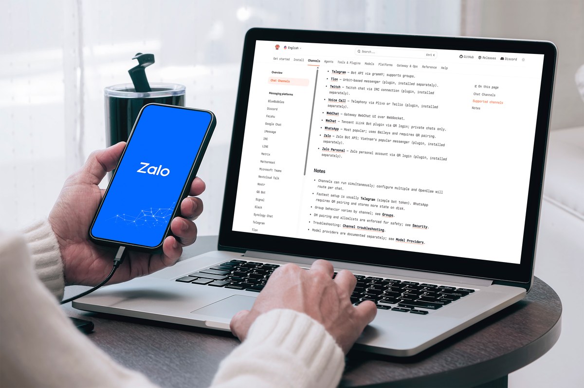 Người dùng gõ laptop bạc, tay cầm điện thoại đen hiển thị Zalo xanh dương trên bàn.